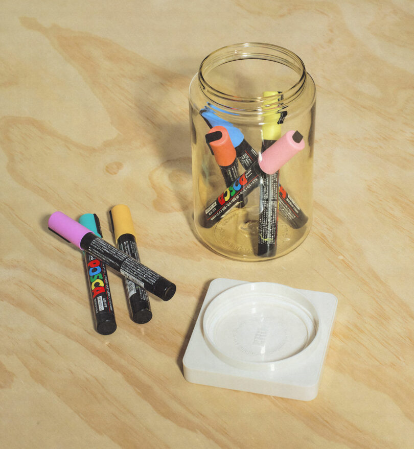 An open glass jar containing colorful markers, with a few markers lying on a wooden surface next to it and a white coaster nearby.