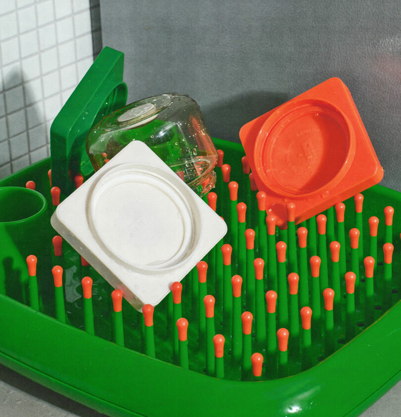 Green drying rack with orange pegs holding a glass bottle, an orange lid, and a white lid in a kitchen setting.