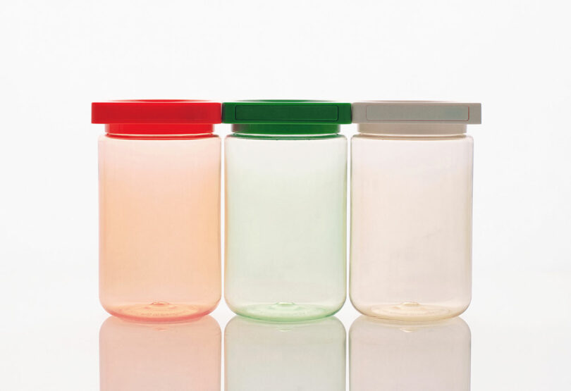 Three empty jars of different transparent colors (pink, green, and clear) with corresponding colored lids (red, green, and gray) are placed side by side against a white background.