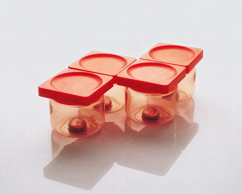Four small transparent plastic containers with red lids are arranged in an L-shape on a white surface.
