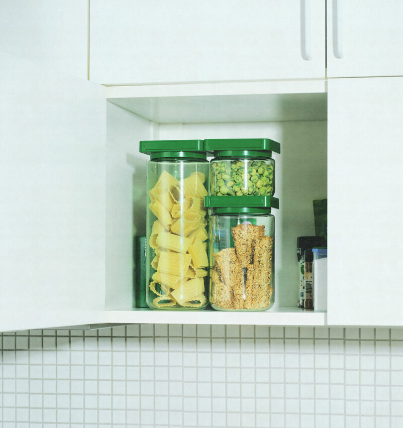 A kitchen cabinet with two shelves holding transparent containers filled with pasta, green peas, and granola bars, all with green lids.