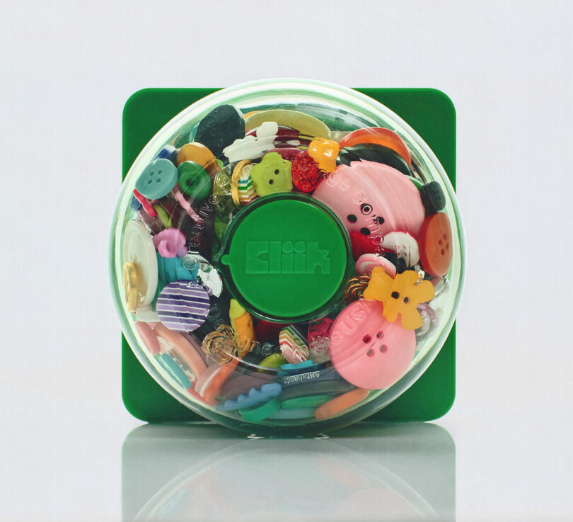A clear round container with a green lid, labeled "Clik", filled with assorted colorful buttons in various sizes and shapes, placed on a reflective surface.