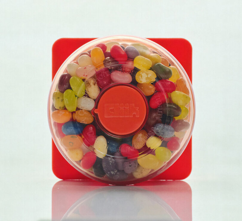 A red dispenser filled with an assortment of colorful jelly beans, encased in a clear plastic cover, is placed on a reflective surface.