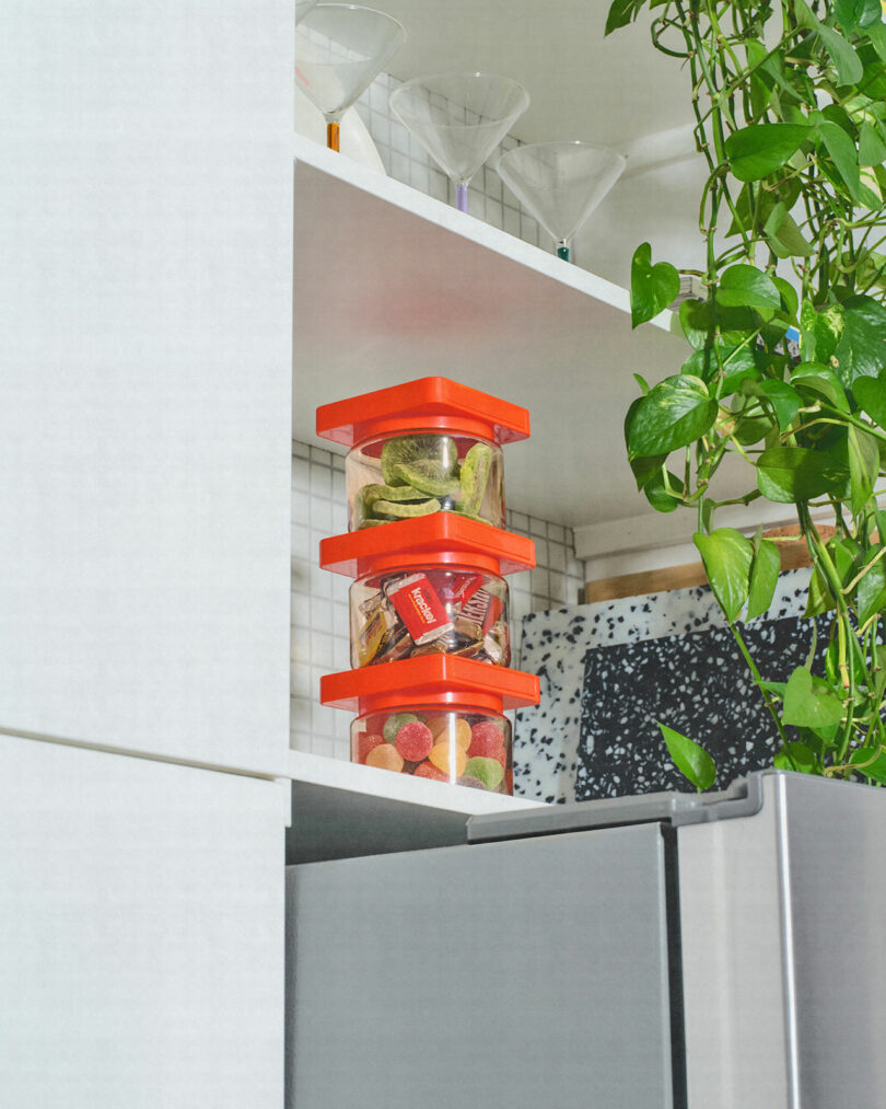 Three red-lidded food storage containers with various snacks are stacked on a kitchen shelf above a refrigerator, next to a plant and martini glasses on an upper shelf.