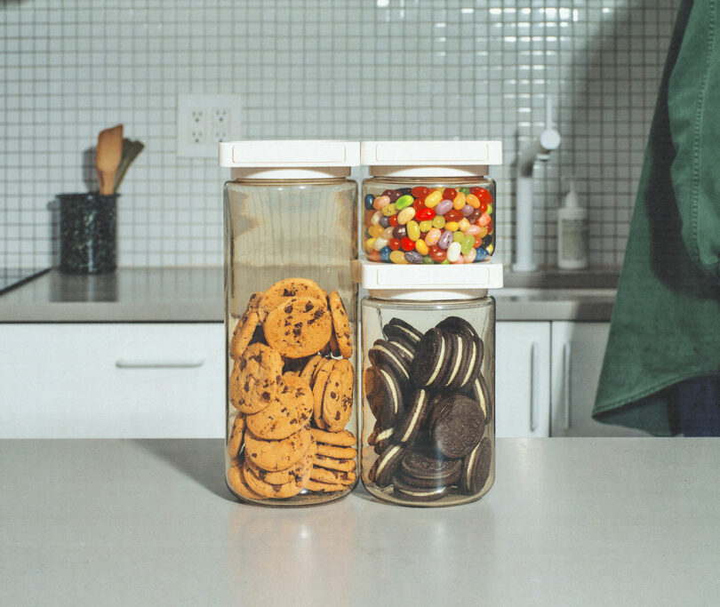 Three transparent jars on a kitchen counter contain chocolate chip cookies, colored jelly beans, and chocolate sandwich cookies. Kitchen utensils and a soap dispenser are visible in the background.