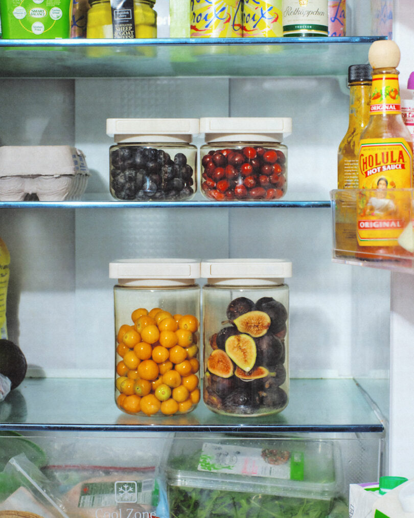 A refrigerator shelf displaying jars of blueberries, cranberries, golden berries, and dried figs, alongside eggs, vegetables, a bottle of hot sauce, and assorted beverages.