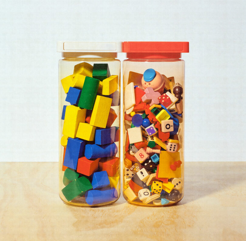 Two clear jars with lids, one containing colorful wooden blocks and the other filled with various small toys, buttons, and knick-knacks, are placed side by side on a wooden surface.