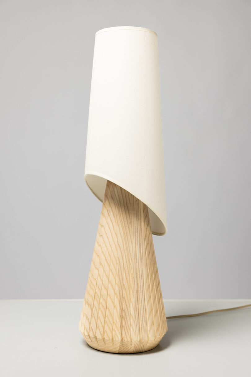 A table lamp with a wooden, cone-shaped base and a white, slightly tilted lampshade placed on a white surface against a light gray background.