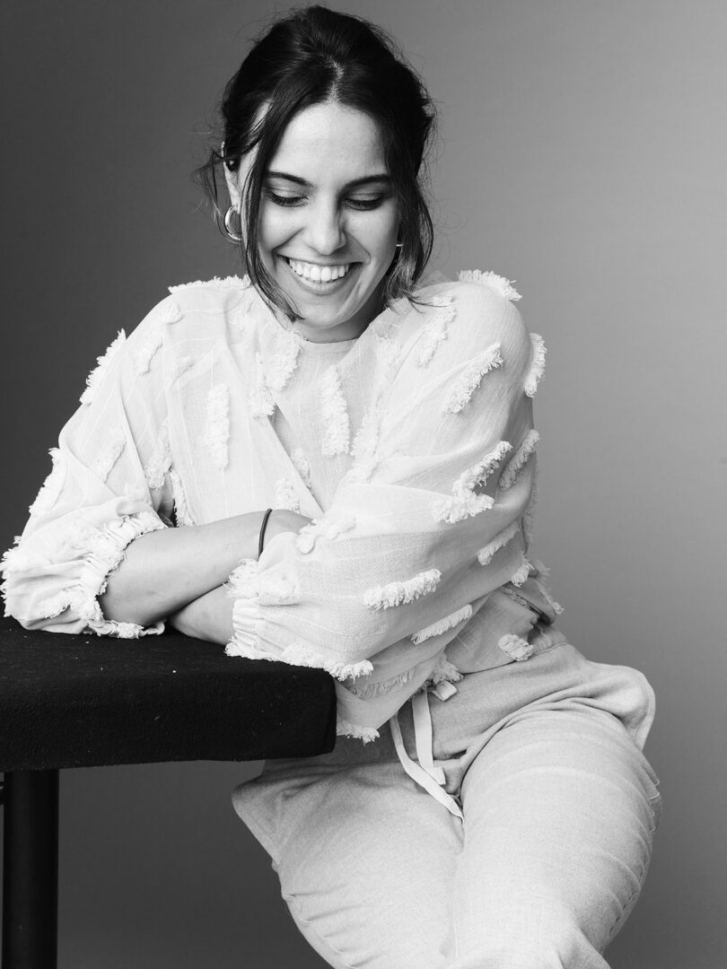 A woman with dark hair tied back, wearing a textured light-colored top and pants, smiles while leaning her elbows on a surface in front of her. The image captures the creative ambiance of designer in black and white.