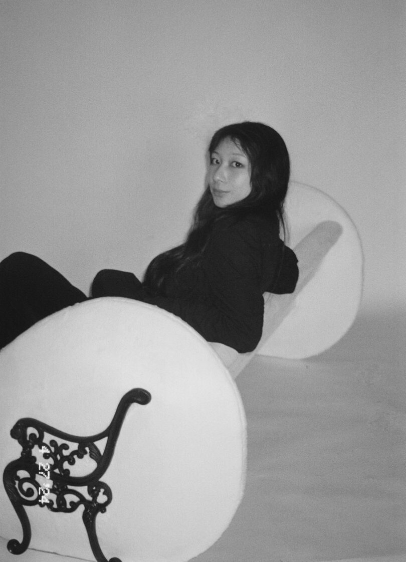 Black and white photo of a person with long hair sitting in a uniquely designed bench with large, round backrest, and decorated armrests.