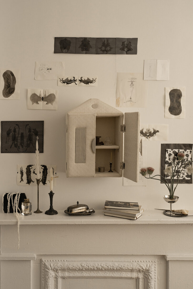 A small cabinet with open doors hangs on a wall, surrounded by various black and white inkblot art pieces. Below it, a white mantel displays candles, books, a small vase with flowers, and other objects.