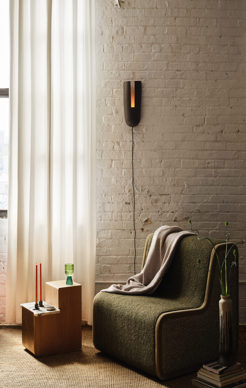 A chair with a blanket faces a small wooden table with red candles and a green glass. A minimalist wall lamp is mounted on a brick wall, next to a sheer curtained window.