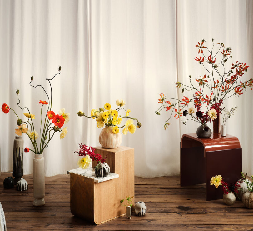 A room with wooden flooring displays various floral arrangements in vases on modern side tables against a backdrop of white curtains.