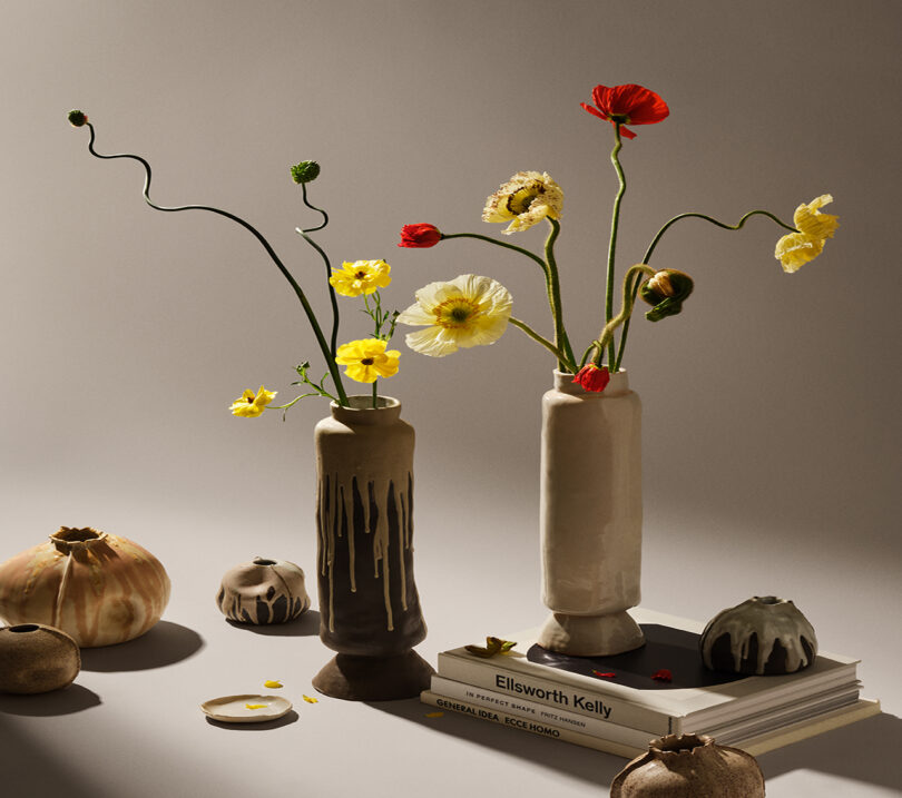 Two vases with uneven, wildflower arrangements sit on a surface with stacked books and decorative, pumpkin-like objects around them. The background is plain and softly lit.