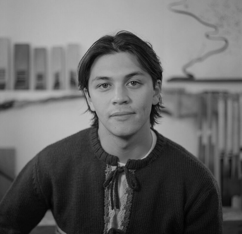 A person with medium-length hair wearing a sweater sits indoors with a neutral expression. Shelves with books and a partially visible abstract artwork are in the background. The image is in black and white.