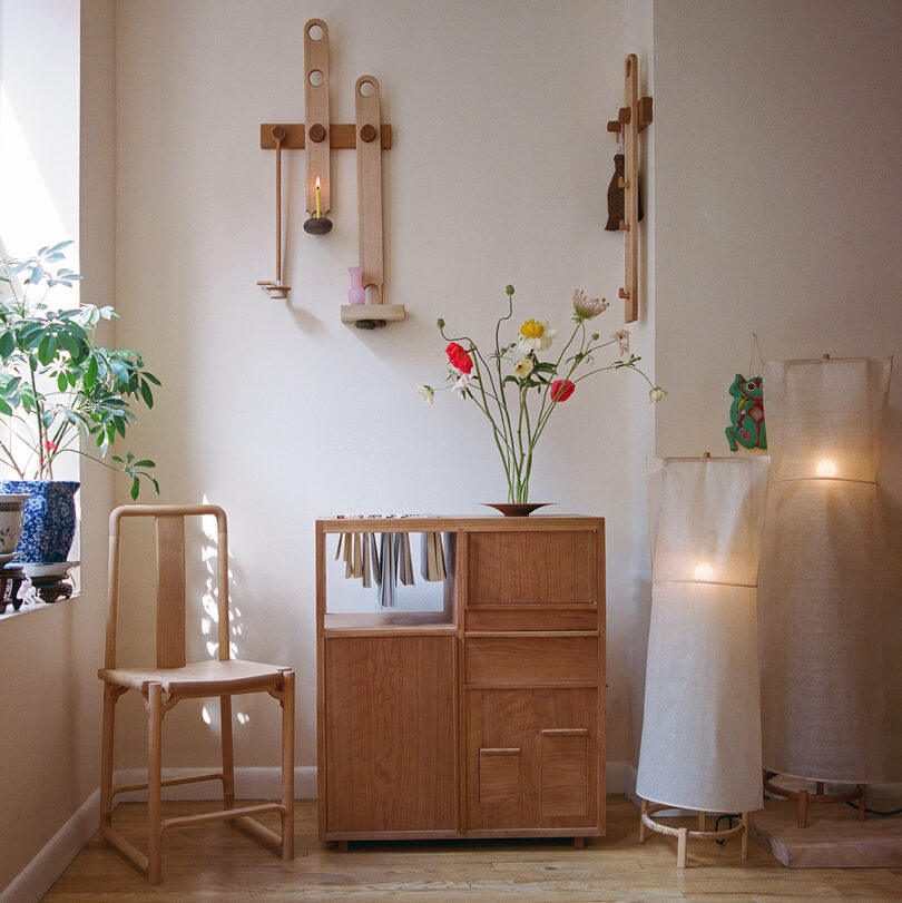 A minimalist room features wooden furniture, a small cabinet, a chair, a lit candle on a wall mount, two floor lamps with fabric shades, and assorted plants and flowers.