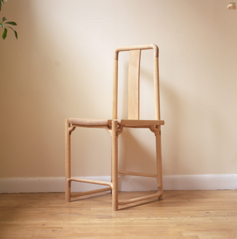 A simple wooden chair with a square frame and straight backrest, placed on a wooden floor against a beige wall.