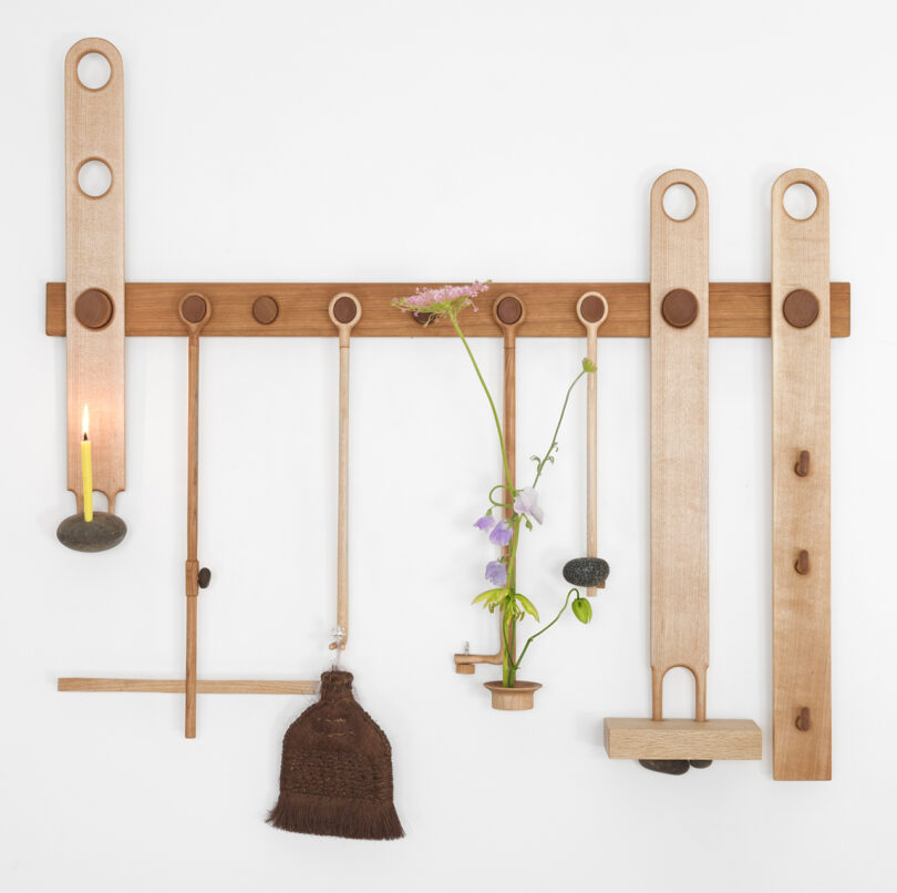 A wooden wall-mounted organizer with hanging items: a lit candle, a small broom, and a vase holding purple flowers.
