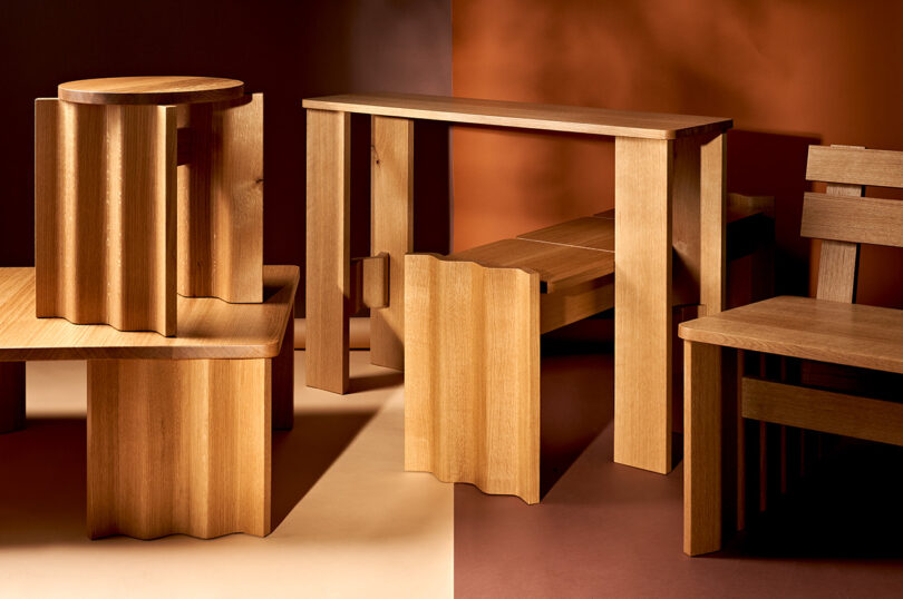 A collection of modern wooden furniture, including stools, a bench, and a table, featuring a design with wavy edges. The setting has a background split into light and dark shades.