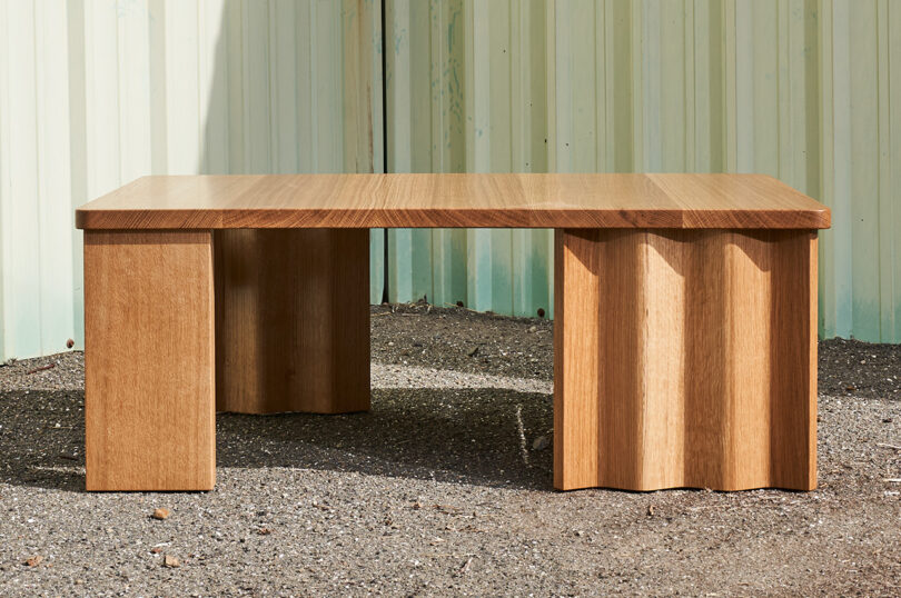 A wooden coffee table with a smooth top and uniquely shaped block legs stands on gravel against a corrugated metal backdrop.