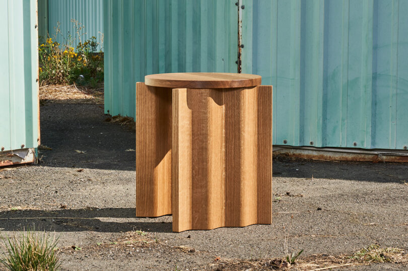 A wooden stool with a round seat and corrugated sides is placed on an outdoor path between two light blue metal structures.