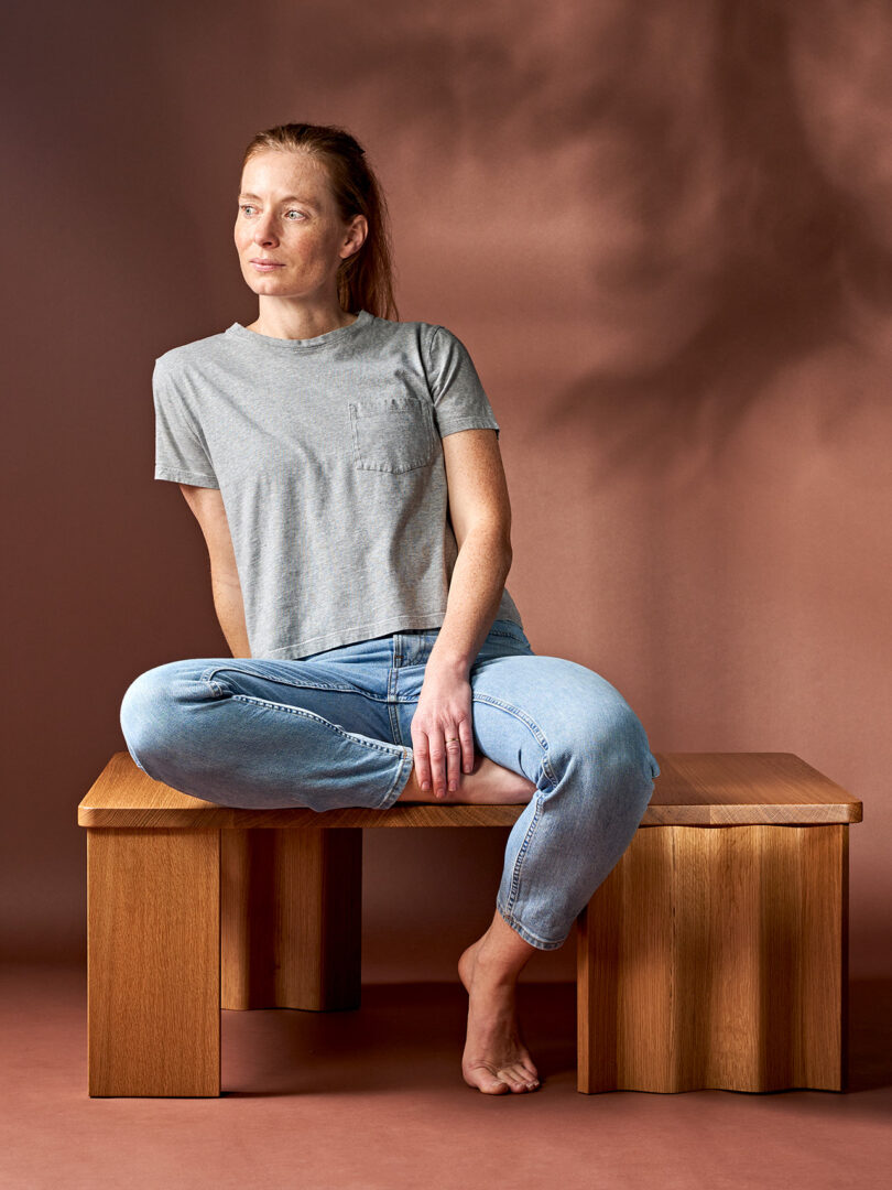 A woman in a gray t-shirt and blue jeans sits barefoot on a wooden bench, looking to her left against a brown background.