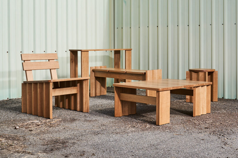 A set of wooden furniture, including a chair, bench, side table, and coffee table, placed on an outdoor gravel surface against a corrugated metal wall.