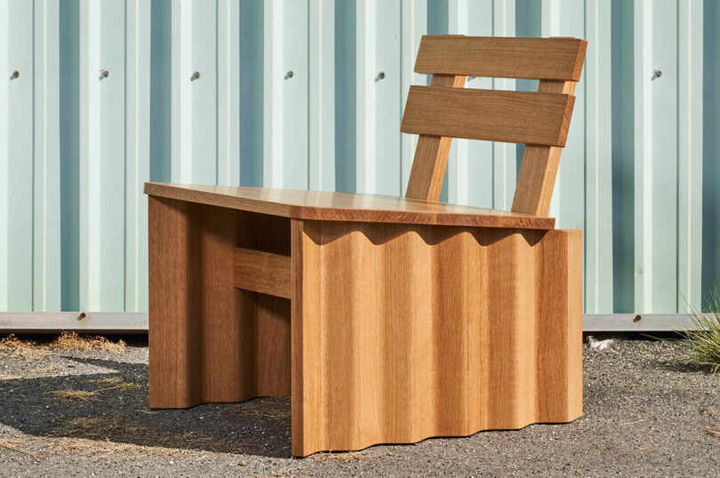 A wooden chair with a unique wavy base design, featuring a slatted backrest, is placed on an outdoor surface in front of a corrugated metal wall.
