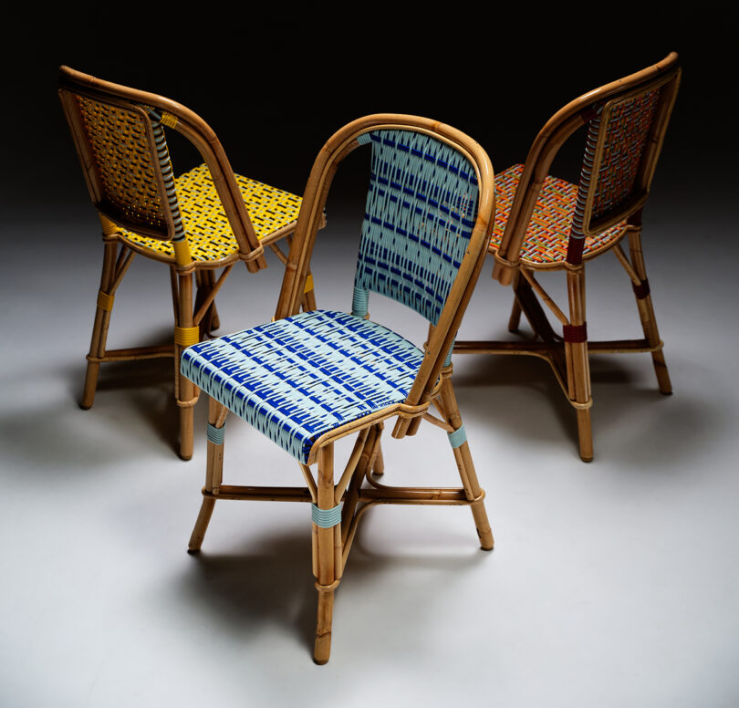 Three chairs with curved backs and woven seats in yellow, blue, and multicolored patterns arranged in a semicircle on a plain background.