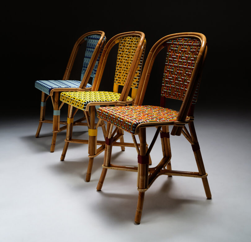 Three wooden chairs with colorful woven seats and backrests are arranged in a row on a gray floor with a dark background.