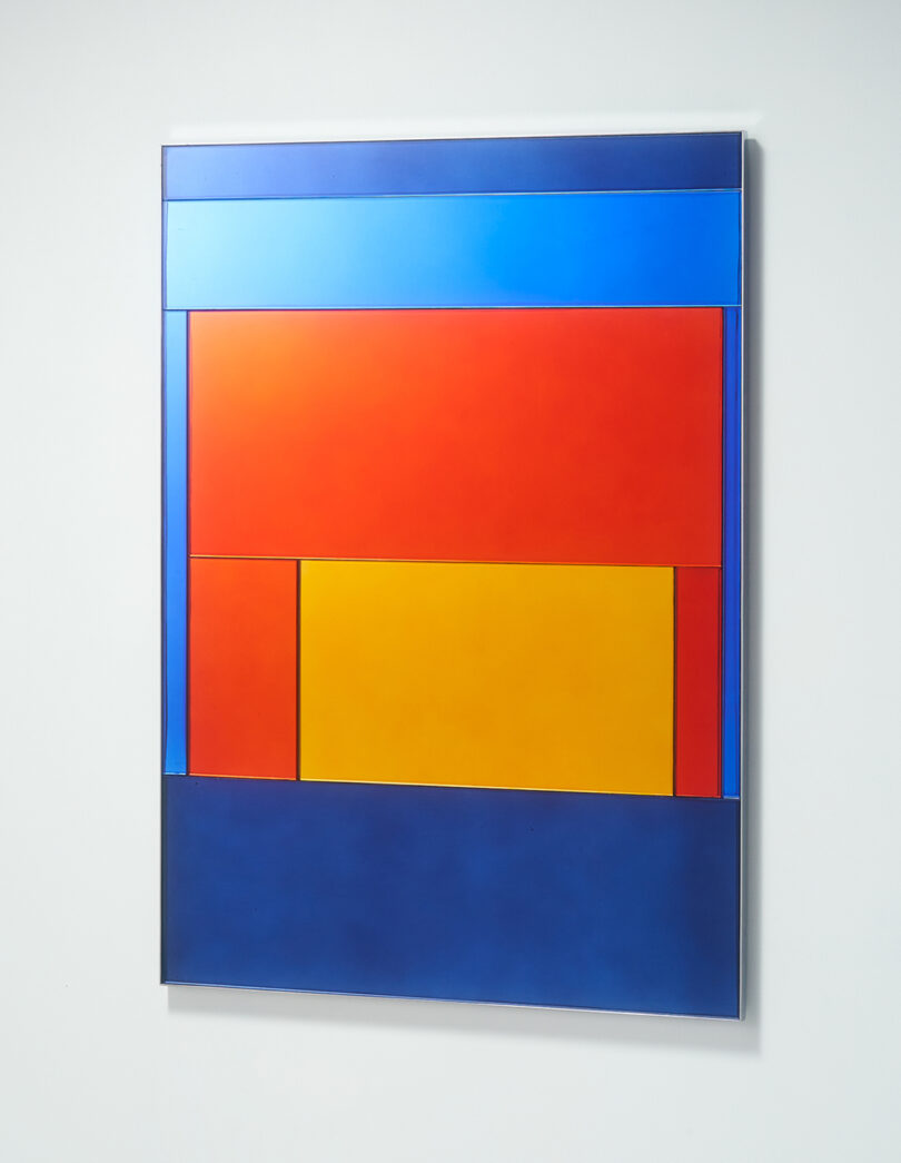 A geometric abstract mirror with rectangles in blue, red, and yellow hues arranged in a symmetrical pattern against a white wall.