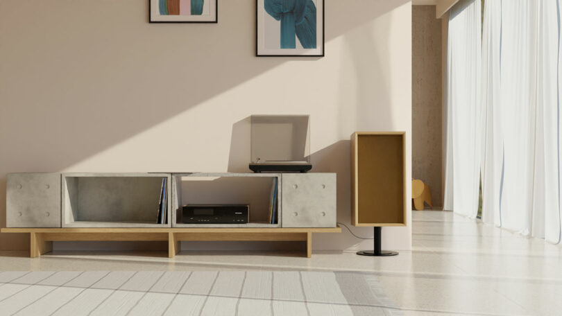 A minimalistic living room features a media console holding vinyl records, a turntable, and a speaker. Two framed artworks are hung on the wall above the console, while sunlight streams through large windows.