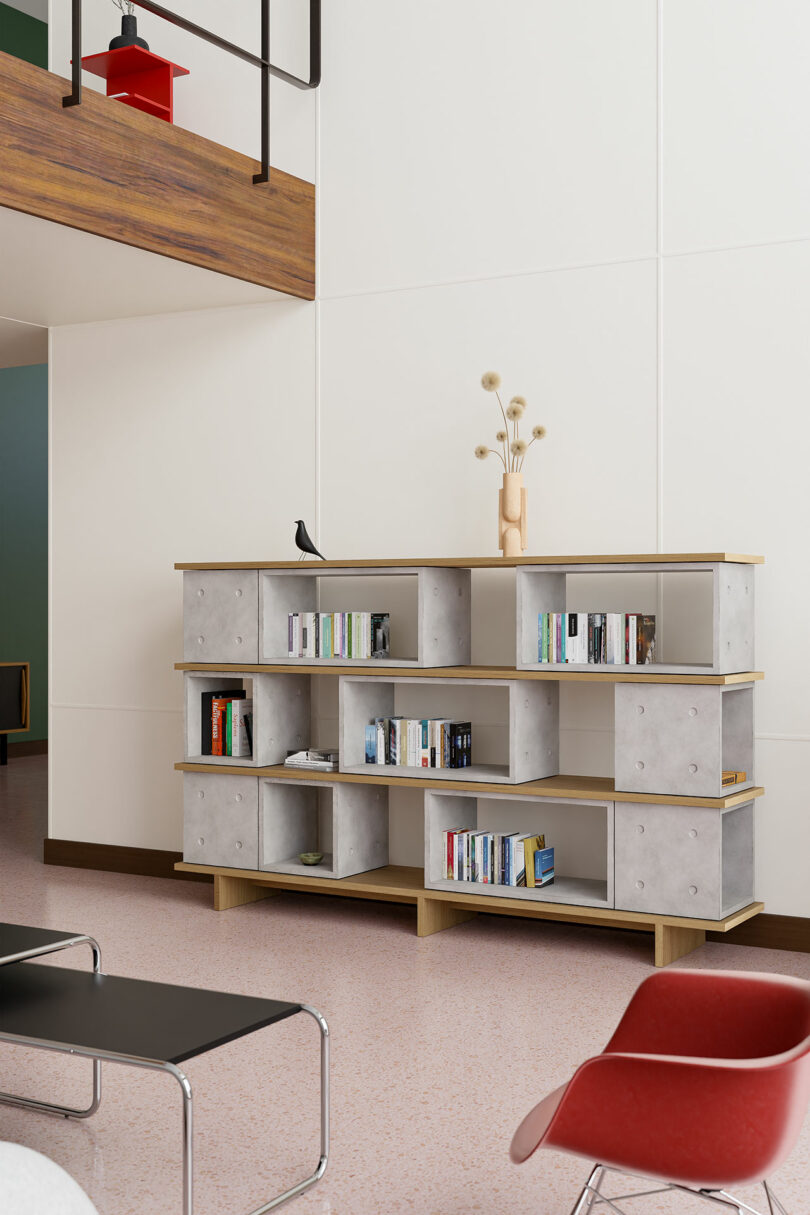 A modern living room features a three-tier bookshelf with various books, decorative objects, and a small bird figurine. The room boasts minimalist furniture including a red chair and a black bench.