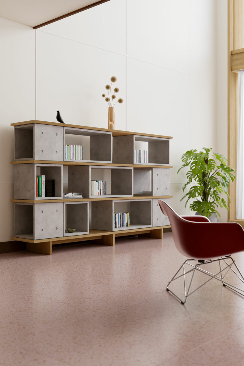 A modern living room with a large bookshelf containing books and decorative items, a potted plant, and a red chair with a wire base on a pink terrazzo floor, complemented by sleek accents.