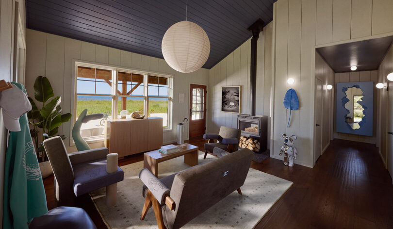 A cozy living room with modern decor, featuring chairs, a sofa, a pendant light, and a wood-burning stove. Beyond the large windows, there's a view of a grassy area and the ocean in the distance.