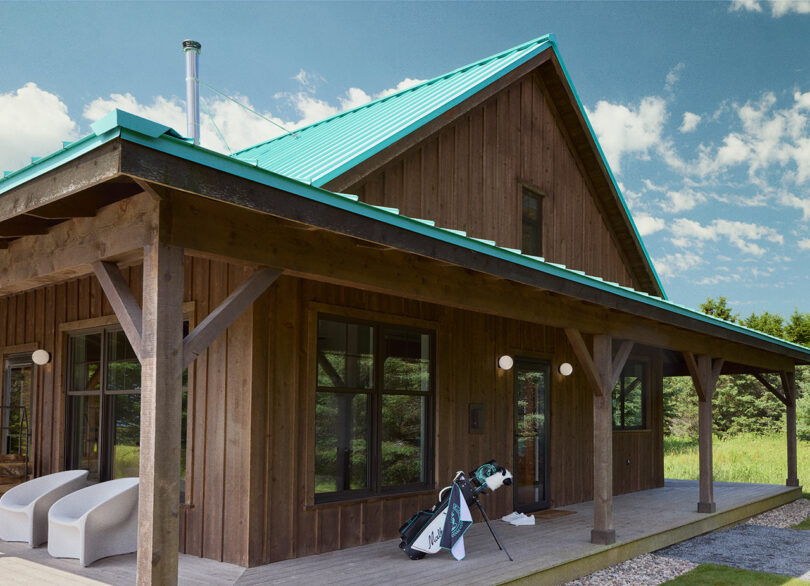Wooden cabin with teal metal roof, large porch, white outdoor chairs, and a golf bag on the porch. Green trees and partly cloudy sky in the background.