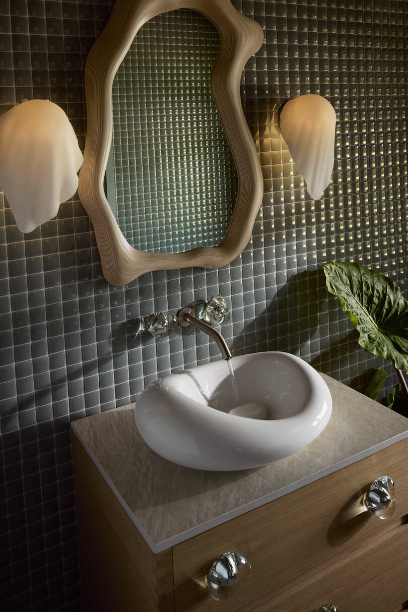 A modern bathroom vanity setup features a uniquely shaped white basin, a wooden framed mirror, and wall sconce lights on a textured tiled wall. A green plant is partially visible on the right.