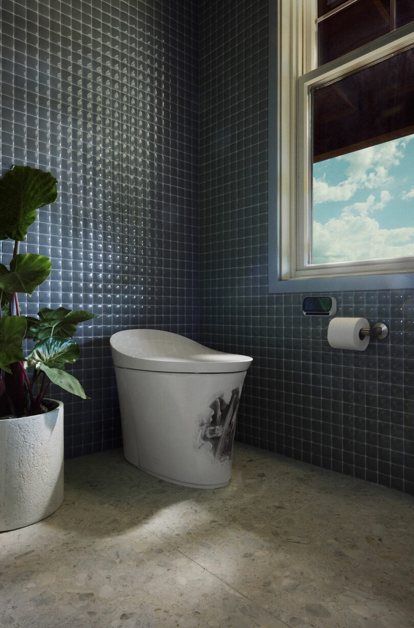 A modern bathroom features a sleek white toilet, a wall-mounted toilet paper holder, a potted plant, and a window providing natural light. The walls are covered in small, dark gray tiles.