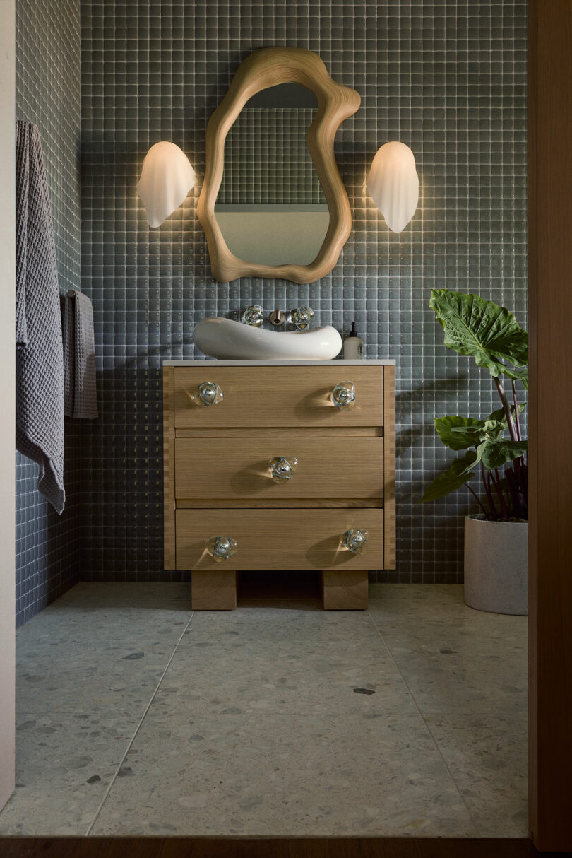 A bathroom with gray tiled walls, a wooden vanity with a wavy mirror, two wall-mounted lights, a white sink, and a potted plant on the right.
