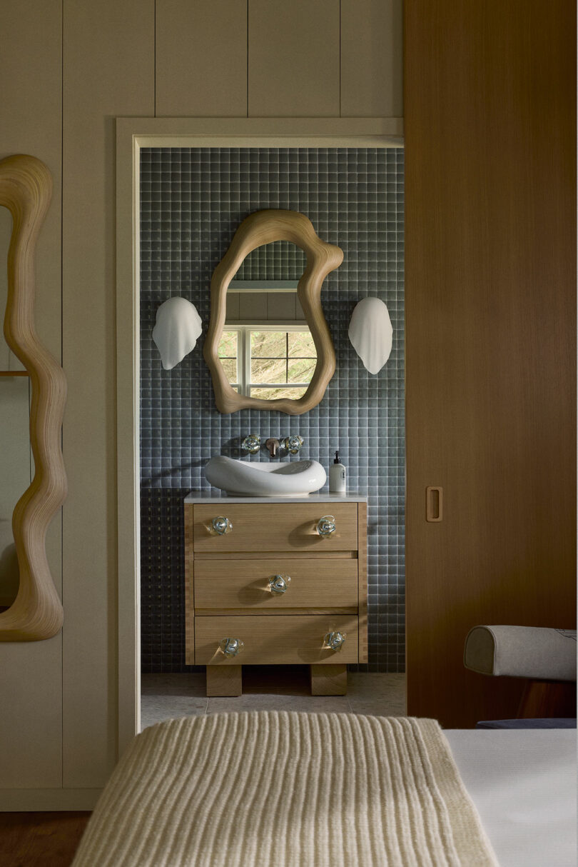 Modern bathroom features a wooden vanity below a wavy mirror, flanked by white wall sconces, and a textured blue tile backsplash. Partial view of a bed with a knitted beige throw in foreground.