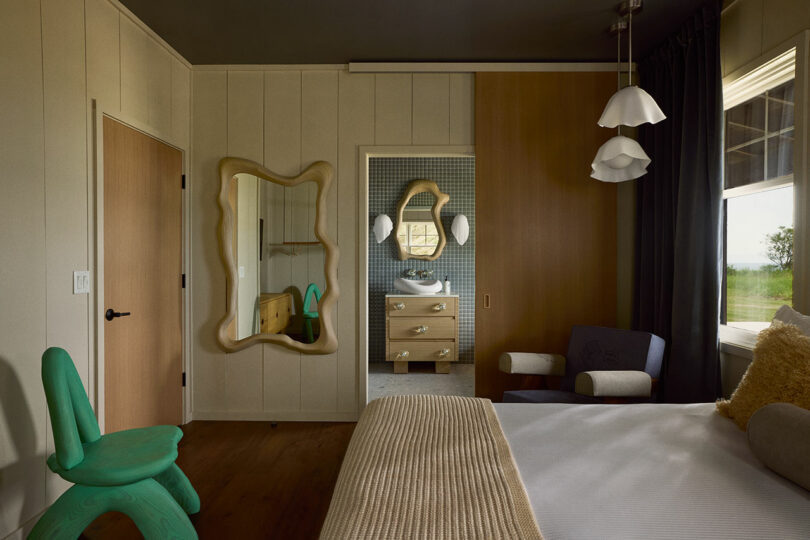 A modern bedroom portrays a mix of minimalism and artistic decor with a unique mirror, green chair, and pendant lights. The room includes a view into a stylish bathroom with a patterned wall and vanity.
