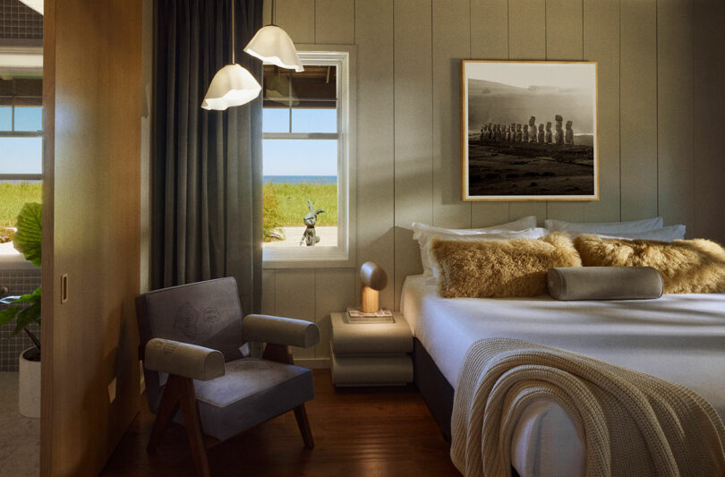 A cozy bedroom features a bed with blankets and fluffy pillows, a unique lamp on the nightstand, a modern chair, and wall art. The window offers a view of the outside landscape.