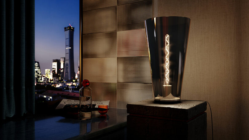 A modern table lamp illuminates a room with a cityscape visible through the window at dusk. A bottle of liquor, a glass, and a magazine are on a nearby table.