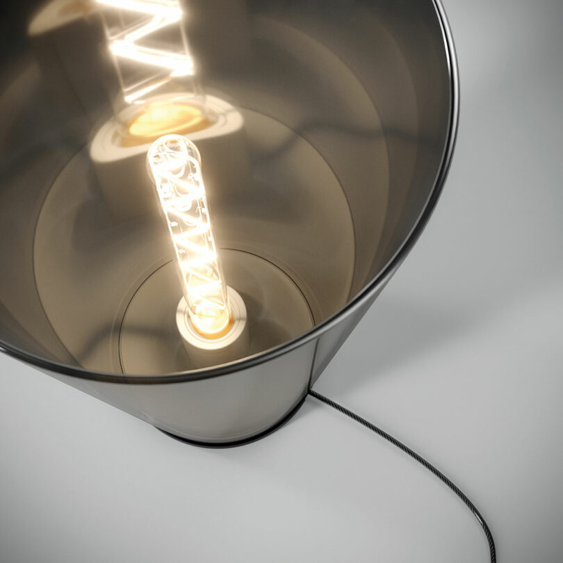 Close-up view of a lit, cylindrical LED filament bulb inside a reflective lamp with a fabric-covered power cord extending from the bottom.