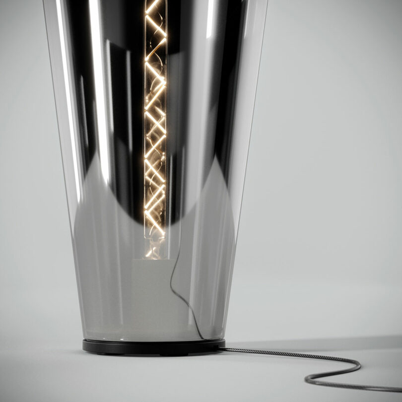 A sleek, modern lamp with a spiral filament light inside a metallic and glass body, connected to a power cord at its base.