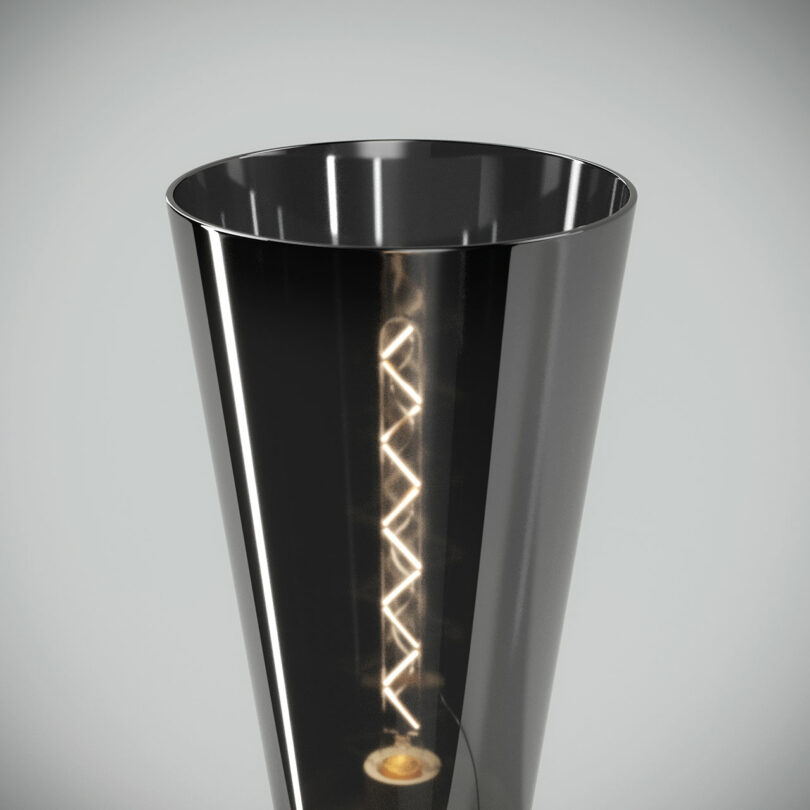 Close-up of a black, conical lamp showing an illuminated spiral filament inside. The lamp has a glossy surface and is placed against a neutral background.
