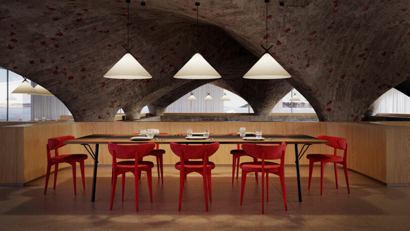 A modern dining area with a long table, red chairs, place settings, and pendant lights. The space has a unique curved ceiling and a warm, inviting atmosphere.