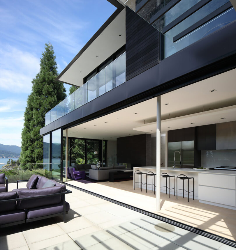 A modern house with large glass doors opening to a spacious patio. The interior features minimalist décor with a kitchen and living area overlooking a leafy landscape and water in the distance.