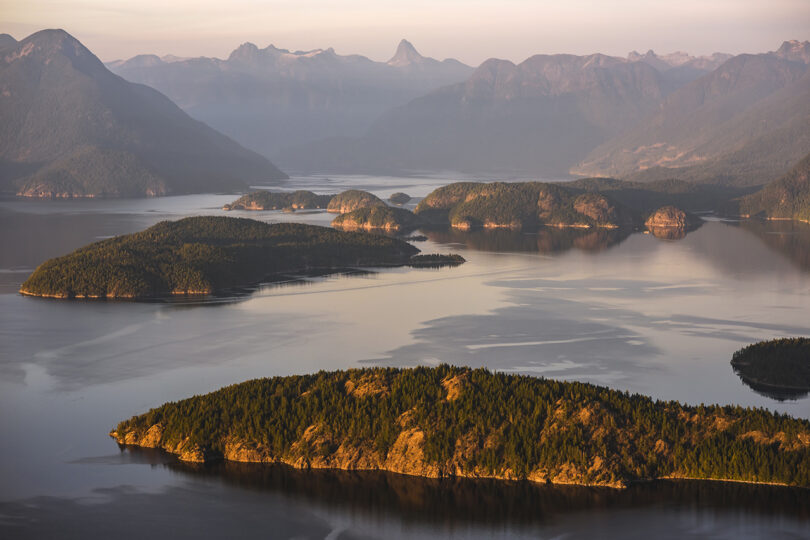 A serene view of several forested islands and mountain ranges in the background, with calm waters reflecting the landscape during a hazy sunset.