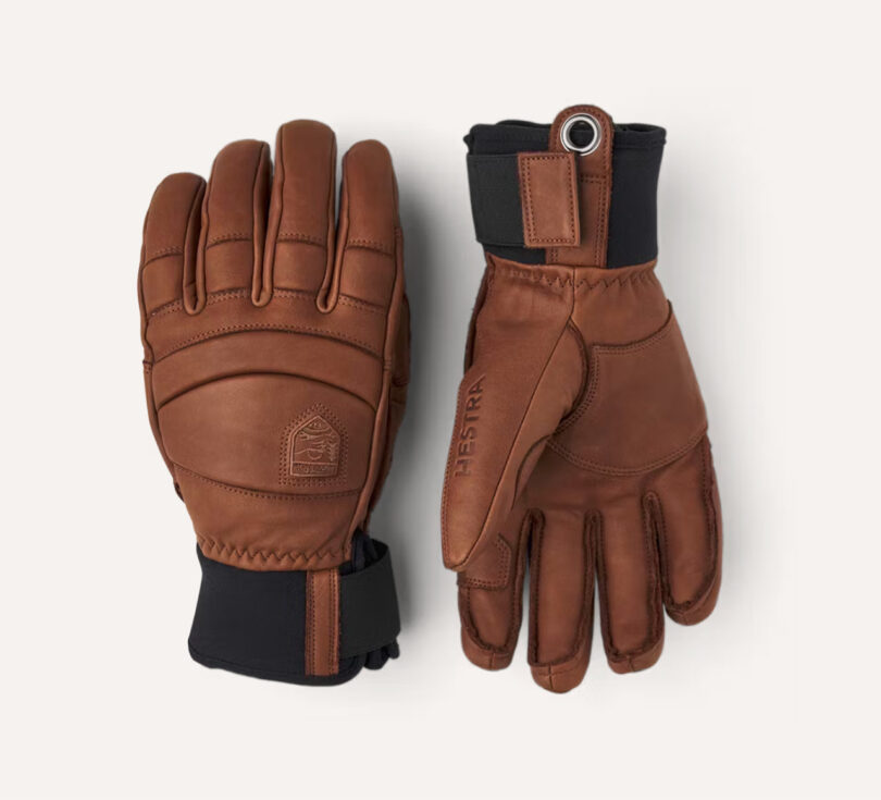A pair of brown leather gloves with black wrist cuffs and the brand "Hestra" embossed on the wrist. One glove is shown palm up and the other palm down.